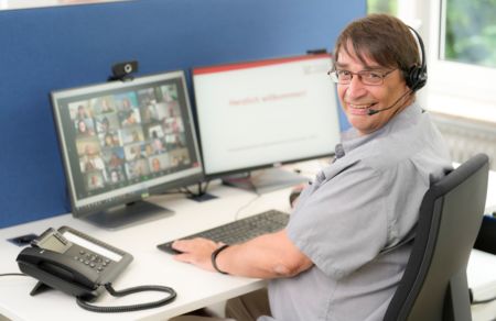 Kollege Rainer Wallbruch sitzt am PC. Er schaut über seine linke Schulter und lächelt in die Kamera. Im Hintergrund erkennt man verschwommen seinen PC-Monitor mit vielen Kacheln und Teilnehmenden von einem Online-Meeting