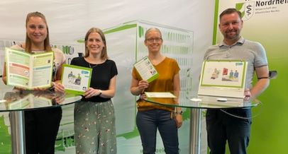 Foto: Das Projektteam steht an zwei Stehttischen. Im Hintergrund ist eine Stellwand mit dem Logo von der Ärztekammer Nordrhein. Das Projektteam hält die Materialien in Leichter Sprache in den Händen und in die Kamera.