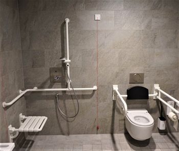 Ein barrierefreier Toilettenraum mit Duschmöglichkeit, Alle Ausstattungslemente sind weiß und heben sich gut gegen die braune Wand ab. Im Duschbereich sind Haltegriffe und ein Klappsitz montiert.