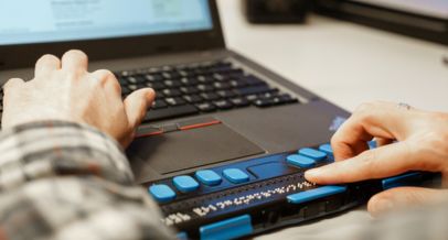 Zwei Hände, die auf einer Braille-Tastatur für den PC liegen. Im Hintergrund, unscharf zu erkennen, der Laptop inklusive Bildschirm.