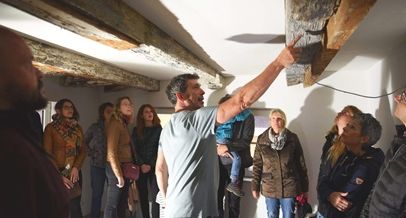 Ein Mann deutet vor einer Gruppe von Menschen in einem niedrigen Raum auf einen alten Balkon über deren Köpfe.