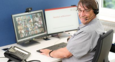 Kollege Rainer Wallbruch sitzt am PC. Er schaut über seine linke Schulter und lächelt in die Kamera. Im Hintergrund erkennt man verschwommen seinen PC-Monitor mit vielen Kacheln und Teilnehmenden von einem Online-Meeting