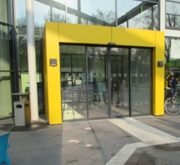 Foto von einem Gebäudezugang. Durch eine breite Portalumrandung in kräftigem Gelb ist der Eingang gut zu erkennen.