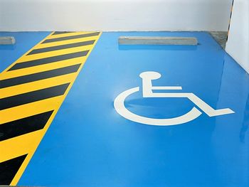 Ein barrierefreier PKW-Stellplatz in einer Parkgarage ist auf dem Boden mit einem großen weißen Rollstuhlsymbol auf kräftig-blauem Grund markiert. Die Bewegungsfläche neben der Stellfläche ist in gelb-schwarz als Sperrfläche gestrichen.