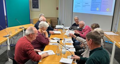 mehrere Menschen sitzen an einem Tisch, vor ihnen liegen die Seminarunterlagen, an der Wand die Beameransicht