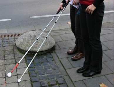 Ein flacher Steinpoller ohne Warnhinweis wird für zwei blinde Frauen mit Langstock zur Stolperfalle.