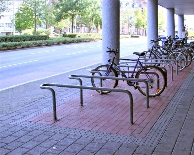 Foto von Fahrradständern als Hindernisse im Bereich eines Gehwegs. Dank eines umlaufenden Aufmerksamkeitstreifens sind die Fahrradständer taktil tastbar.