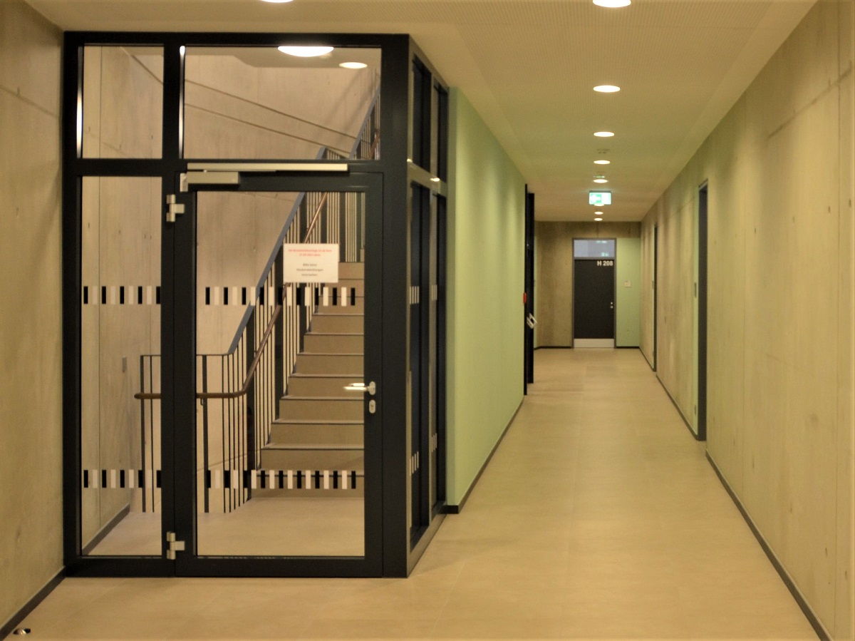 Foto von einem Büroflur mit Blick ins seitliche Treppenhaus. Der Zugang zum Treppenhaus erfolgt durch eine Glastür, die durch schwarze und weiße Glasmarkierungen erkennbar ist.