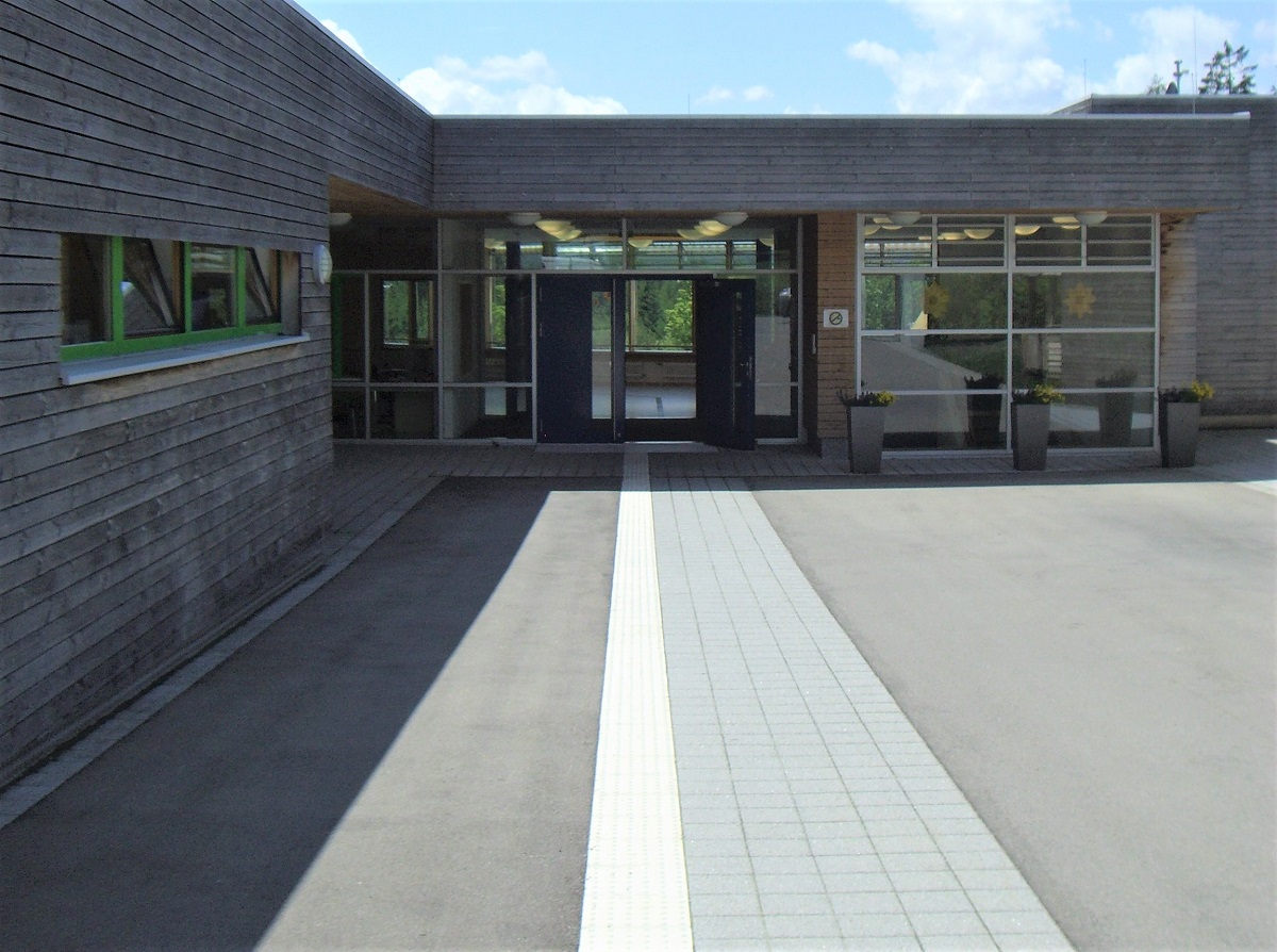 Foto von einem kontrastreichen Leitstreifen aus weißen Rippenplatten neben grauen Gehwegplatten, die auf eine Eingangstür hinführen.