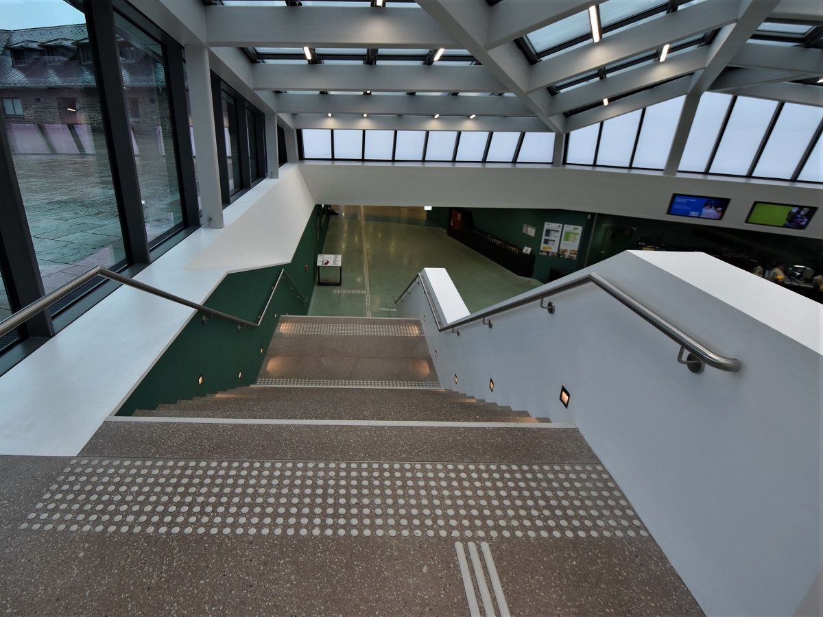 Foto einer Treppe vom Eingang eines Museums zum Infobereich im Untergeschoss. Die Treppe ist gesichert durch kontrastreiche Aufmerksamkeitsfelder und Stufenmarkierungen.