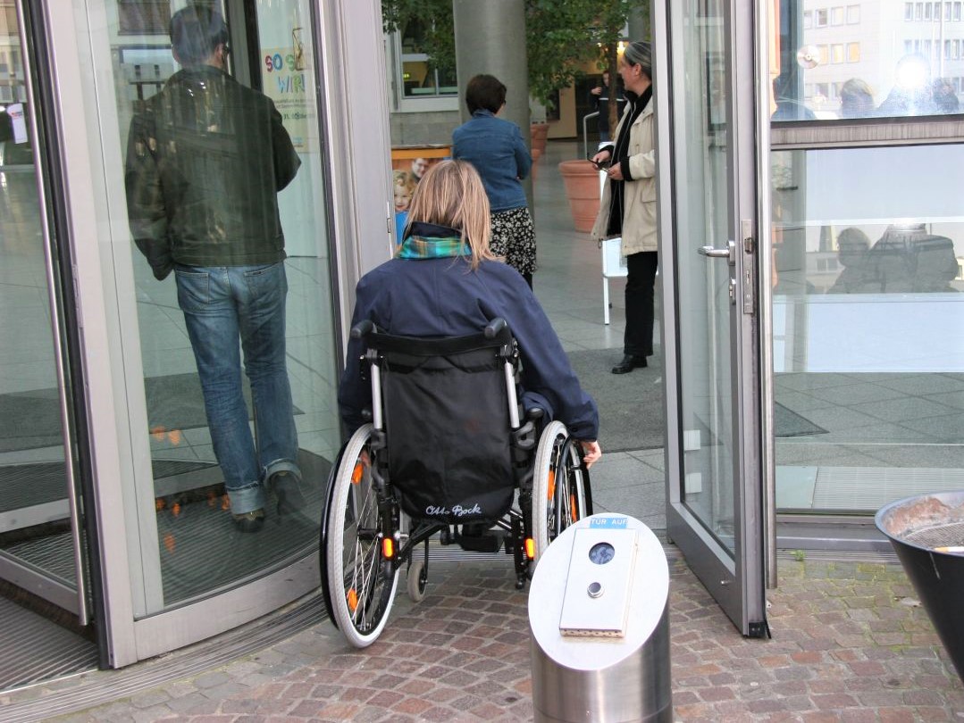 Foto einer Karusselltür mit einer daneben liegenden Automatiktür, durch die eine Rollstuhlfahrerin fährt. Im Vordergrund sieht man den auffälligen Taster für die Automatiktür.